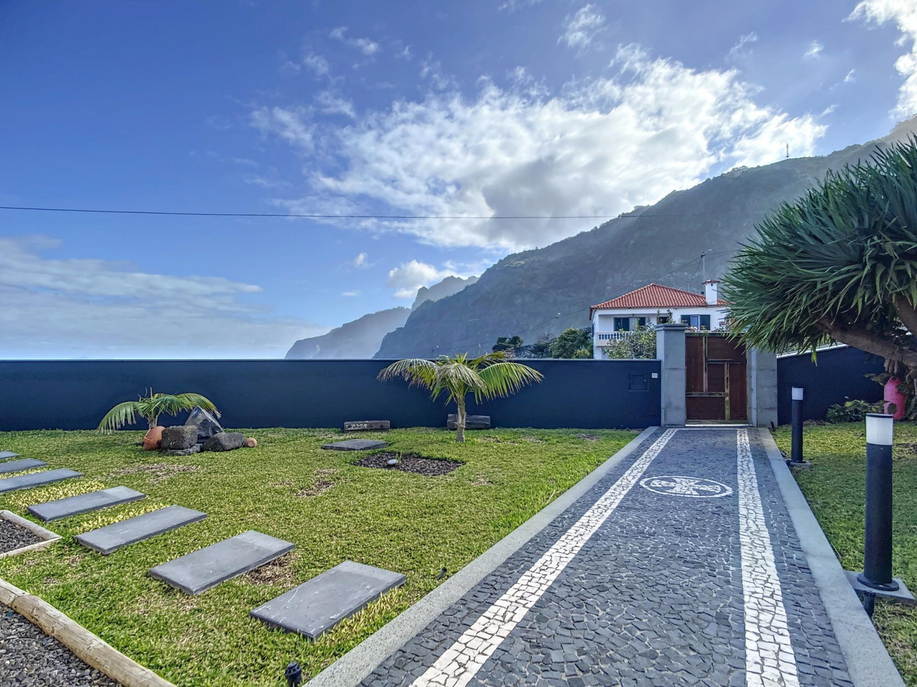 A pathway leading to a house with a red-tiled roof, surrounded by a grassy area with palm trees and a mountain in the background.