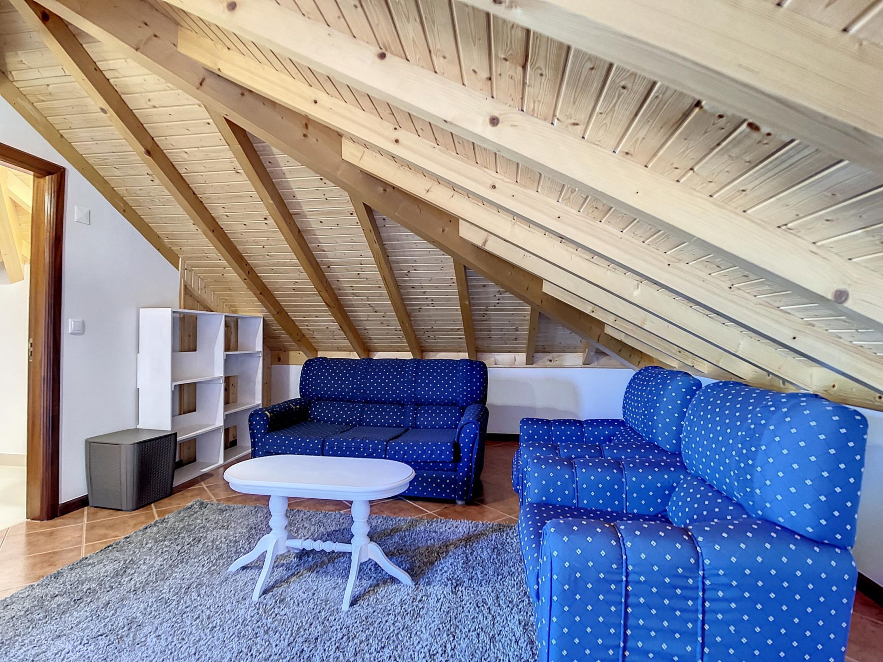 A cozy attic room with a blue sofa set, a white table, and wooden beams on the ceiling.