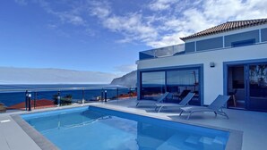 Un área de piscina moderna con agua azul cristalina, sillones de descanso y vistas al océano.
