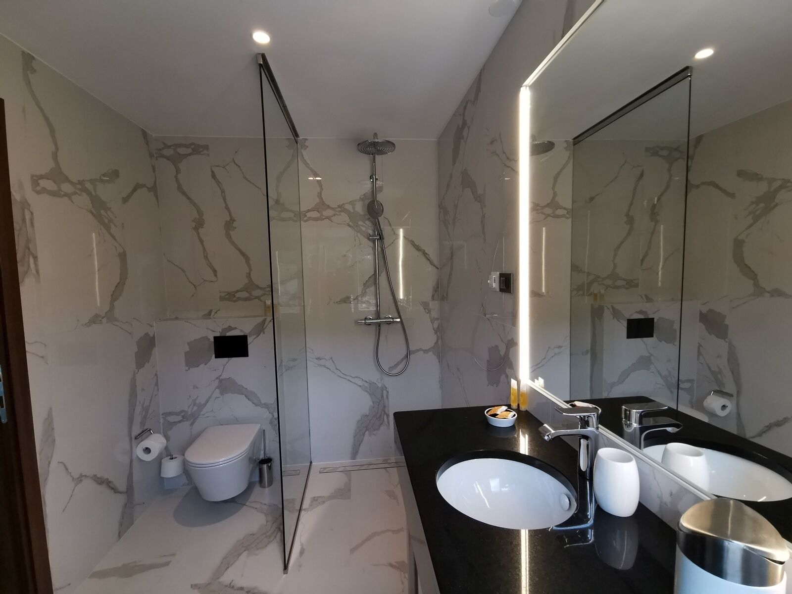 Een moderne badkamer met een douche in een glazen cabine, een wit toilet, een zwarte wastafel met spiegel en marmeren wanden.