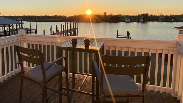 Terraza de madera con dos sillas y una mesa, con vista a una masa de agua tranquila al atardecer.