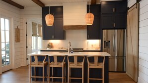 Een keuken met donkere kastjes, een groot eiland met barkrukken, een roestvrijstalen koelkast en hangende rieten hanglampen.