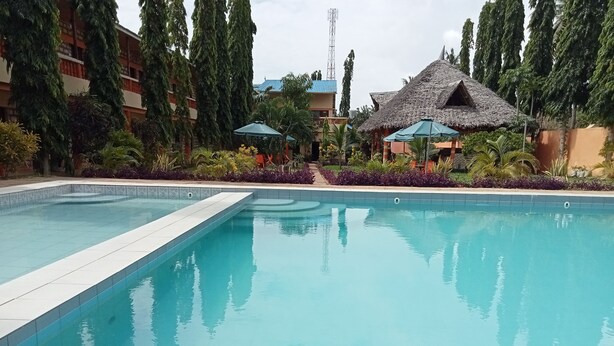 A swimming pool with a tiled edge, surrounded by lush greenery and a thatched-roof structure. - MODERN SOUTH HOTEL (Diani Beach)