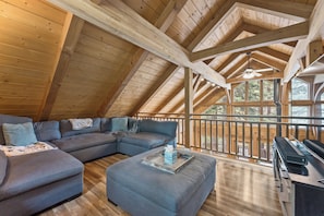 A cozy loft-style room with a large gray sectional sofa, a small ottoman, and a wooden desk with a printer. The room features a sloped ceiling with exposed beams and a large window offering a view of the outdoors.