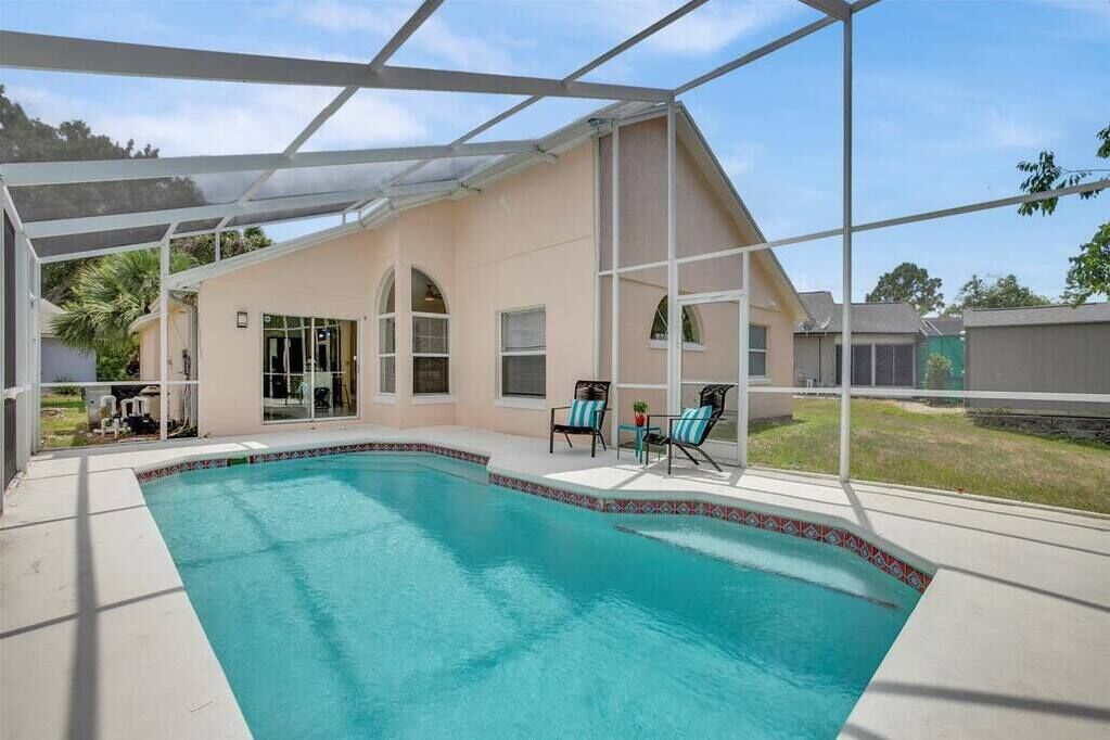 A covered swimming pool with a patio area, chairs, and a view of the surrounding neighborhood.