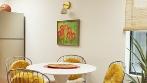 A dining area with a white table and metal chairs, a painting on the wall, and a window with a plant. - Bright, Playful 2bd `Atami Suite` @ La Dea (Santa Fe)
