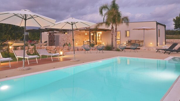 Une piscine avec des chaises longues et des parasols, un mur en pierre et un bâtiment moderne doté d’une terrasse.