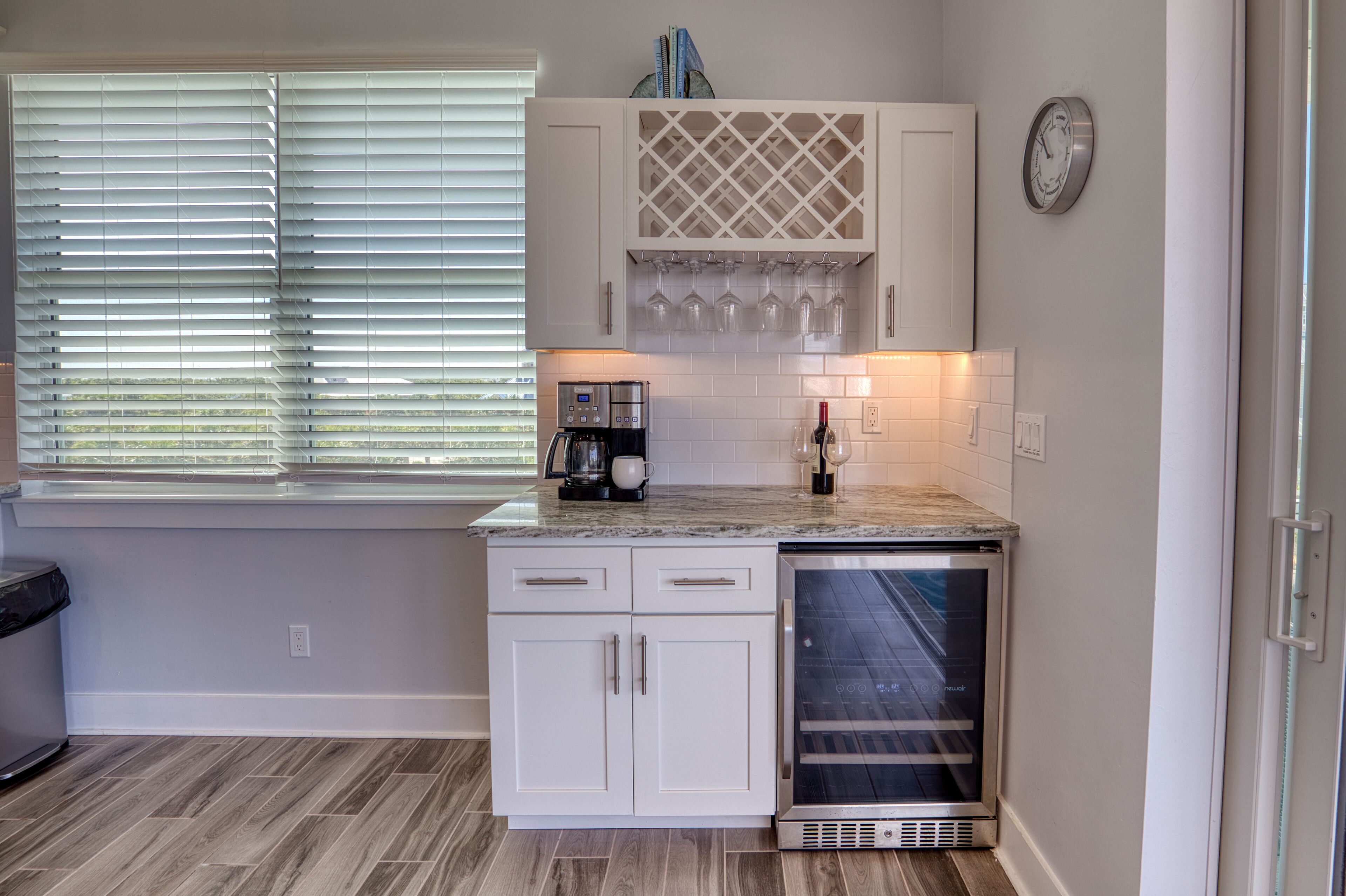 A kitchen with white cabinets, a built-in wine cooler, a coffee maker, and a window with blinds.