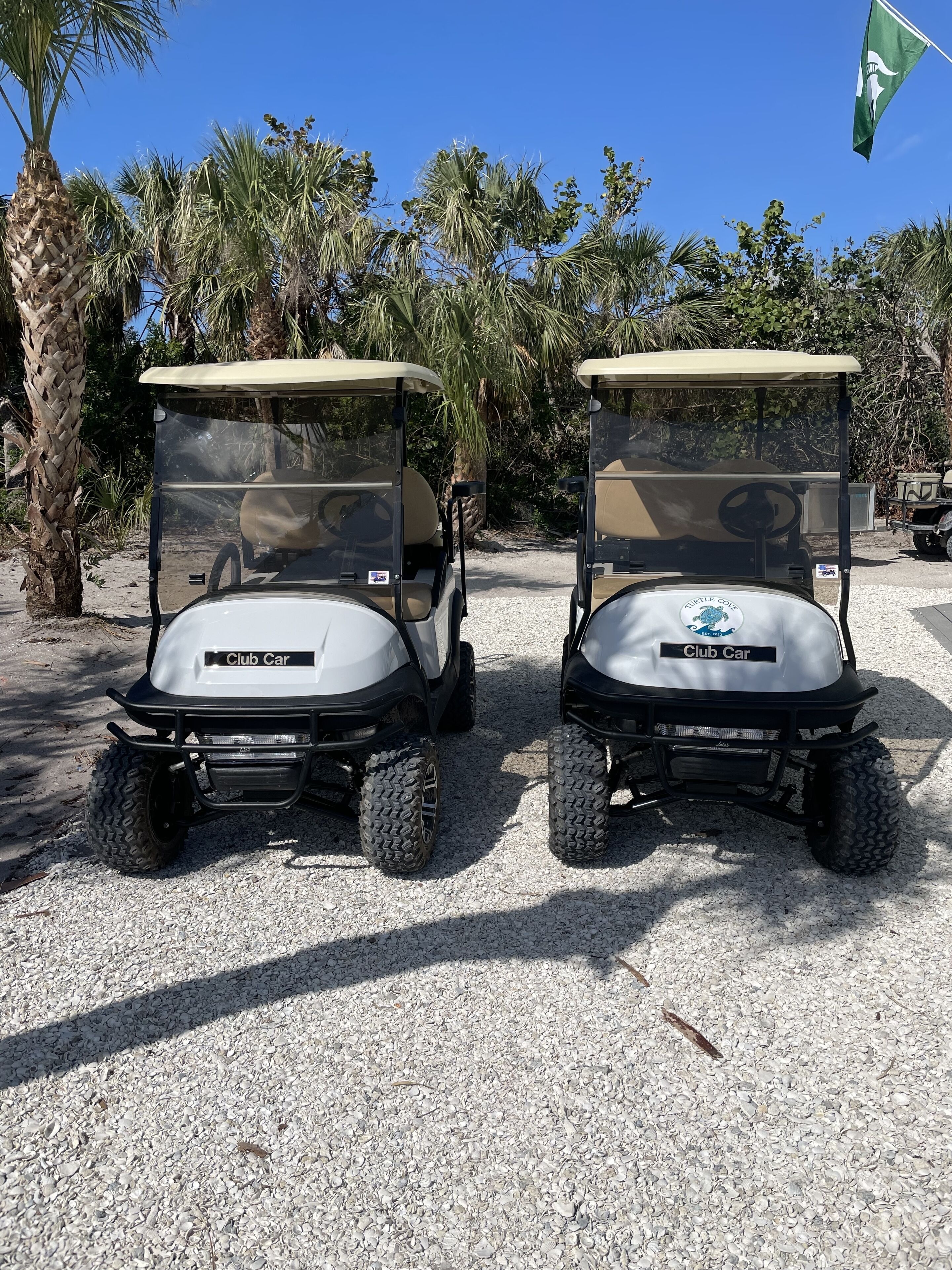 Two golf carts labeled "Club Car" parked on a gravel surface with palm trees and a flag in the background.