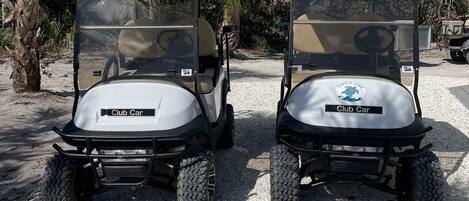 Two golf carts labeled "Club Car" parked on a gravel surface with palm trees and a flag in the background.