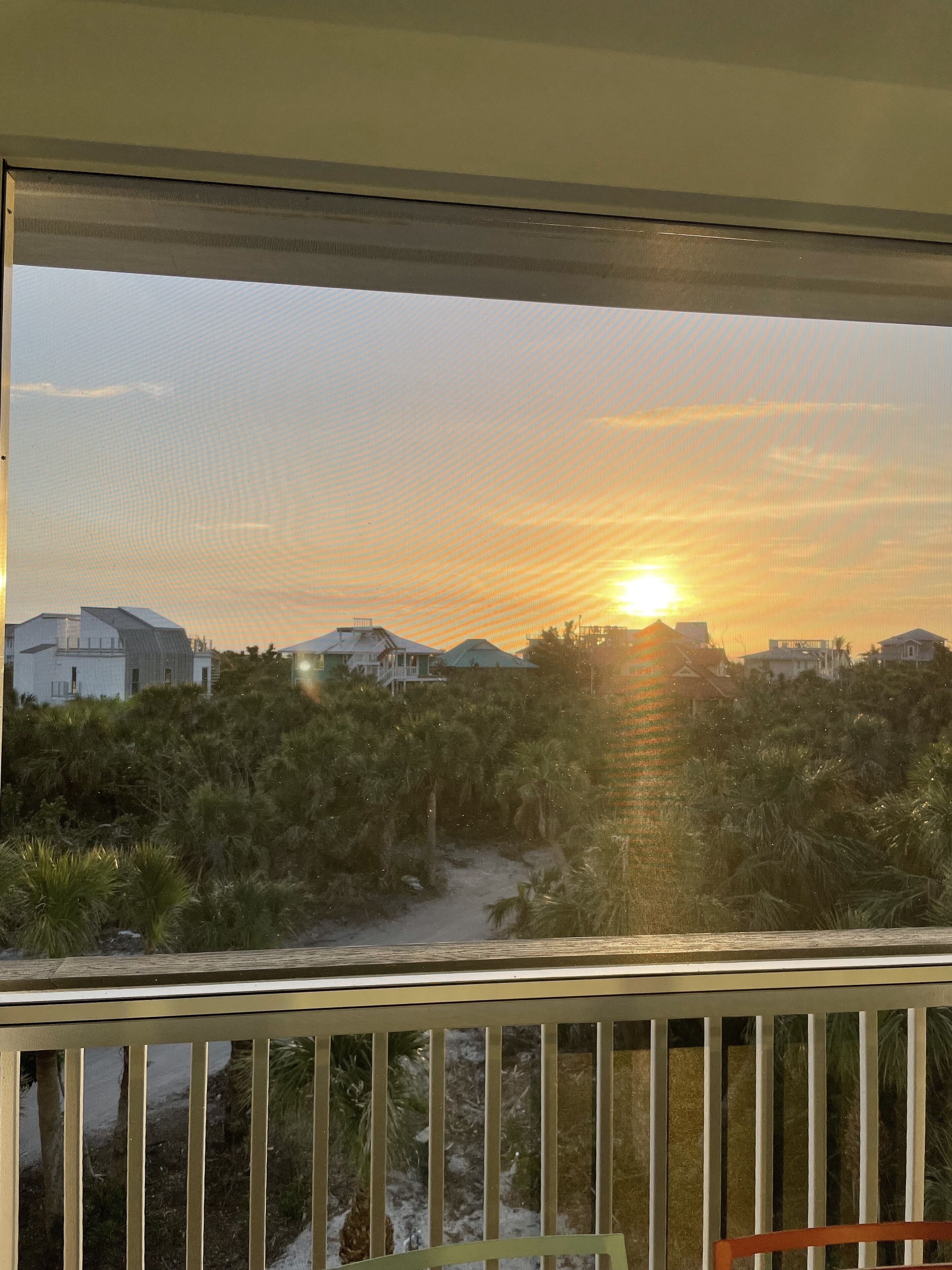Une vue depuis un balcon donnant sur un coucher de soleil, avec des maisons et des palmiers en arrière-plan.