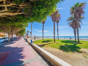A coastal walkway lined with palm trees and parked cars, leading to a beach with people and a playground.