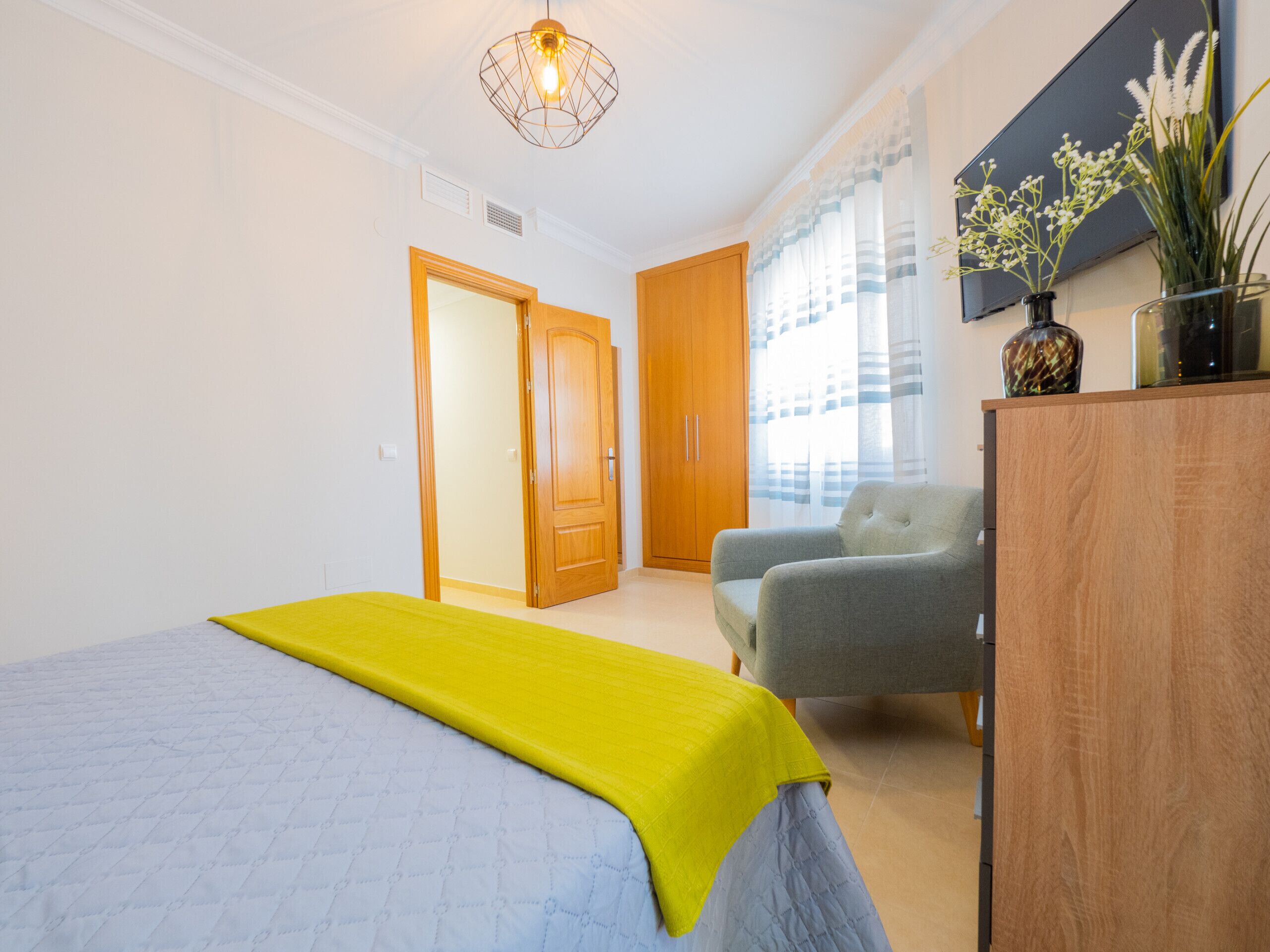 Un dormitorio con cama, una silla, un armario y una ventana con cortinas.