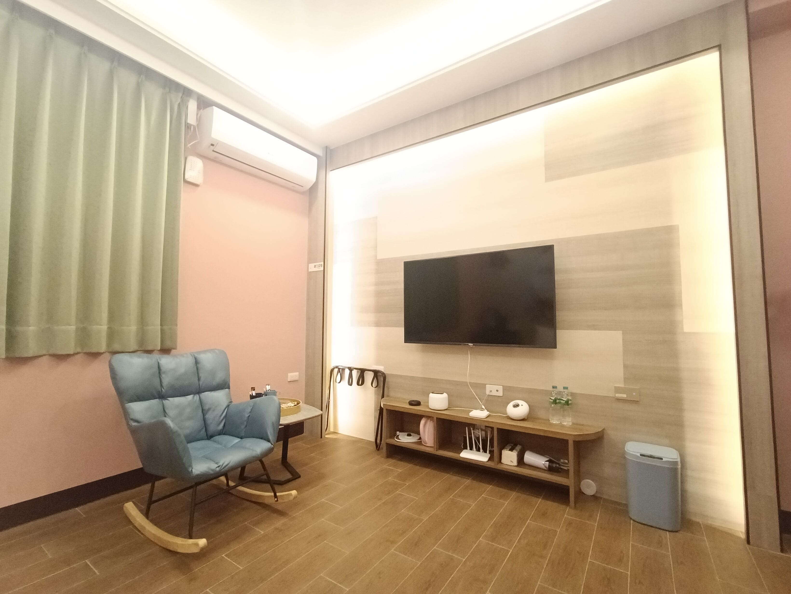 Una sala moderna con un televisor de pantalla plana montado en la pared, un sillón mecedor azul, una mesita con un cuenco y un cesto de basura.