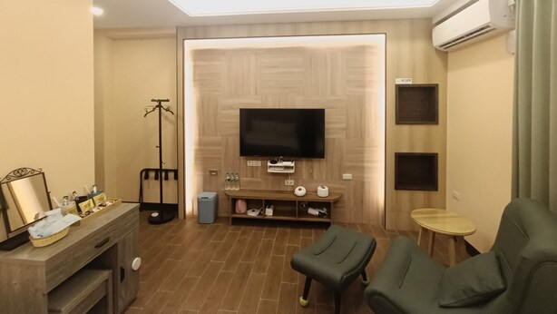 Une chambre d’hôtel moderne avec un téléviseur à écran plat fixé à un mur en bois, une petite table d’appoint en bois, un canapé vert et un lampadaire.