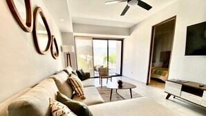 Una sala moderna con un sofá, una mesa de centro y un escritorio. Hay un ventilador de techo, un ventanal con vistas y una puerta que da a otra habitación.
