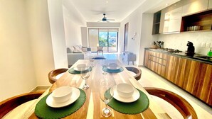 Un comedor moderno con una mesa de madera dispuesta para cuatro personas, una cocina con armarios de madera y vistas al exterior a través de un ventanal corredero.