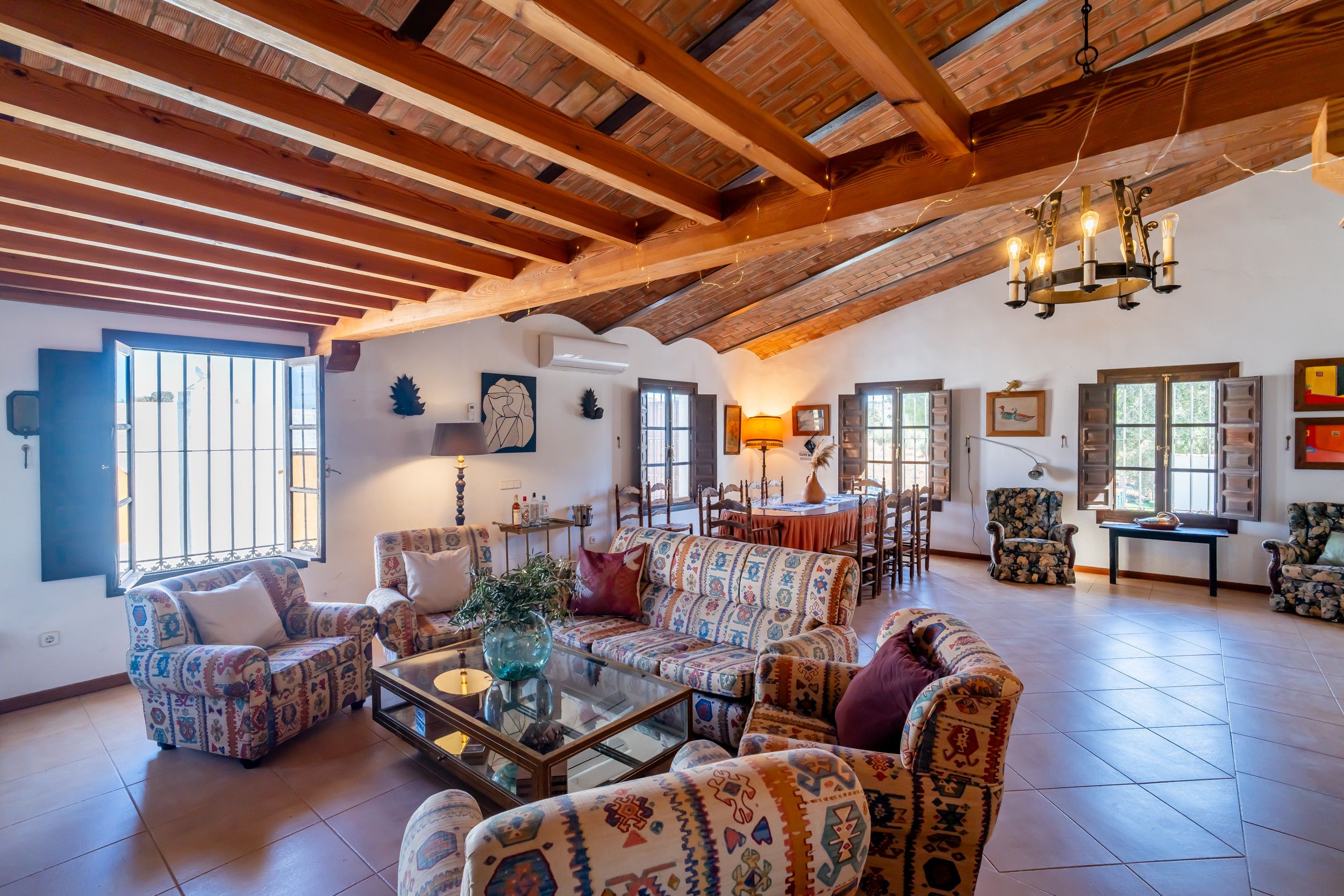 Un vaste espace de vie doté de hauts plafonds et de poutres apparentes, aménagé avec des canapés et des fauteuils à motifs, une table basse en verre et un coin repas comprenant une table et des chaises.