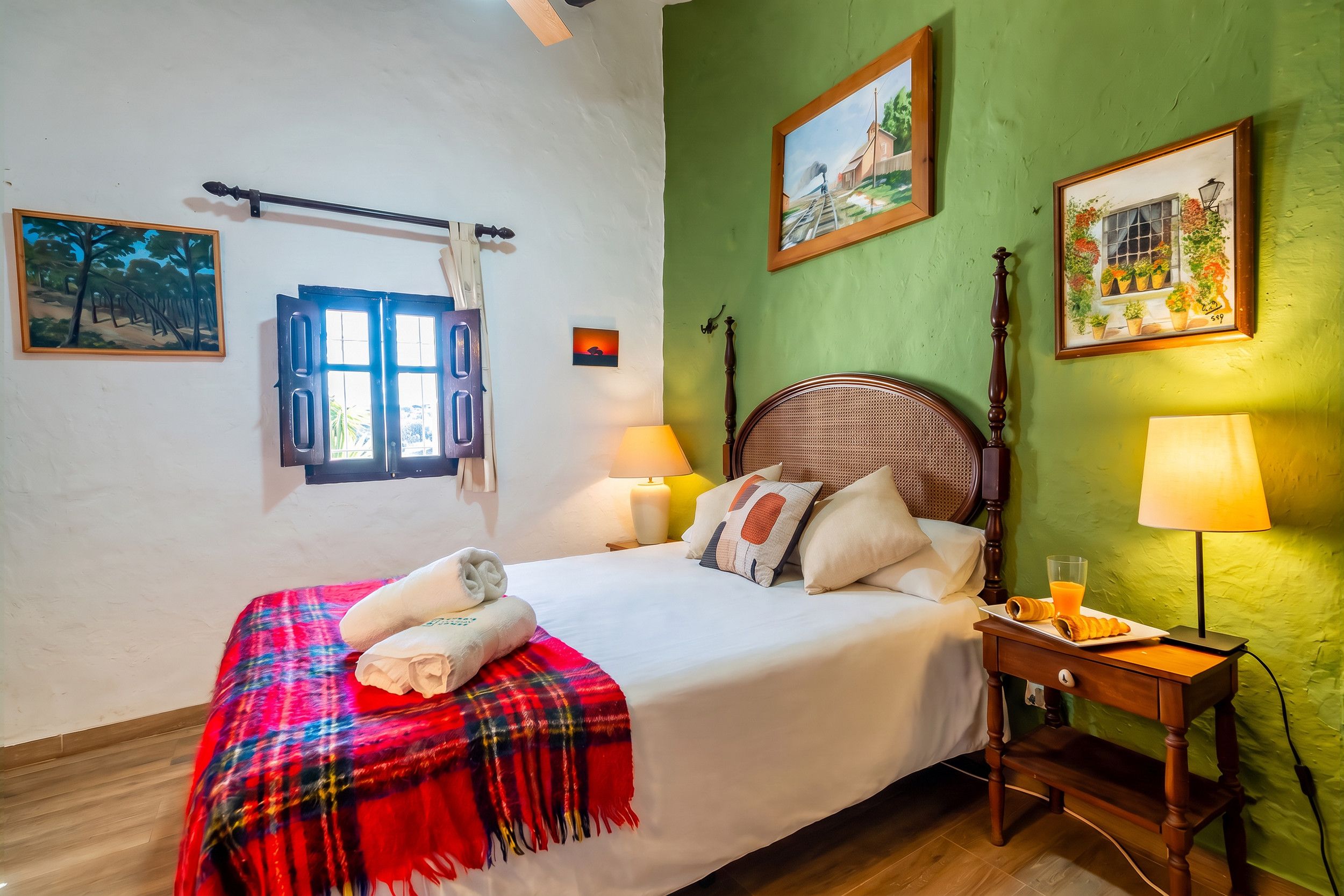 Een hotelkamer met een bed, twee bedlampen, een raam met gordijnen, een nachtkastje met croissants en een glas sinaasappelsap, en twee schilderijen aan de muur.