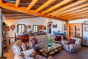 A spacious living room with a high wooden ceiling, patterned sofas, a glass coffee table, and a dining area with chairs and a table.