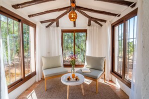 Une pièce chaleureuse avec un plafond en bois, deux fauteuils confortables, une petite table sur laquelle se trouve un livre et un verre de jus d’orange, et une fen être donnant sur les arbres.