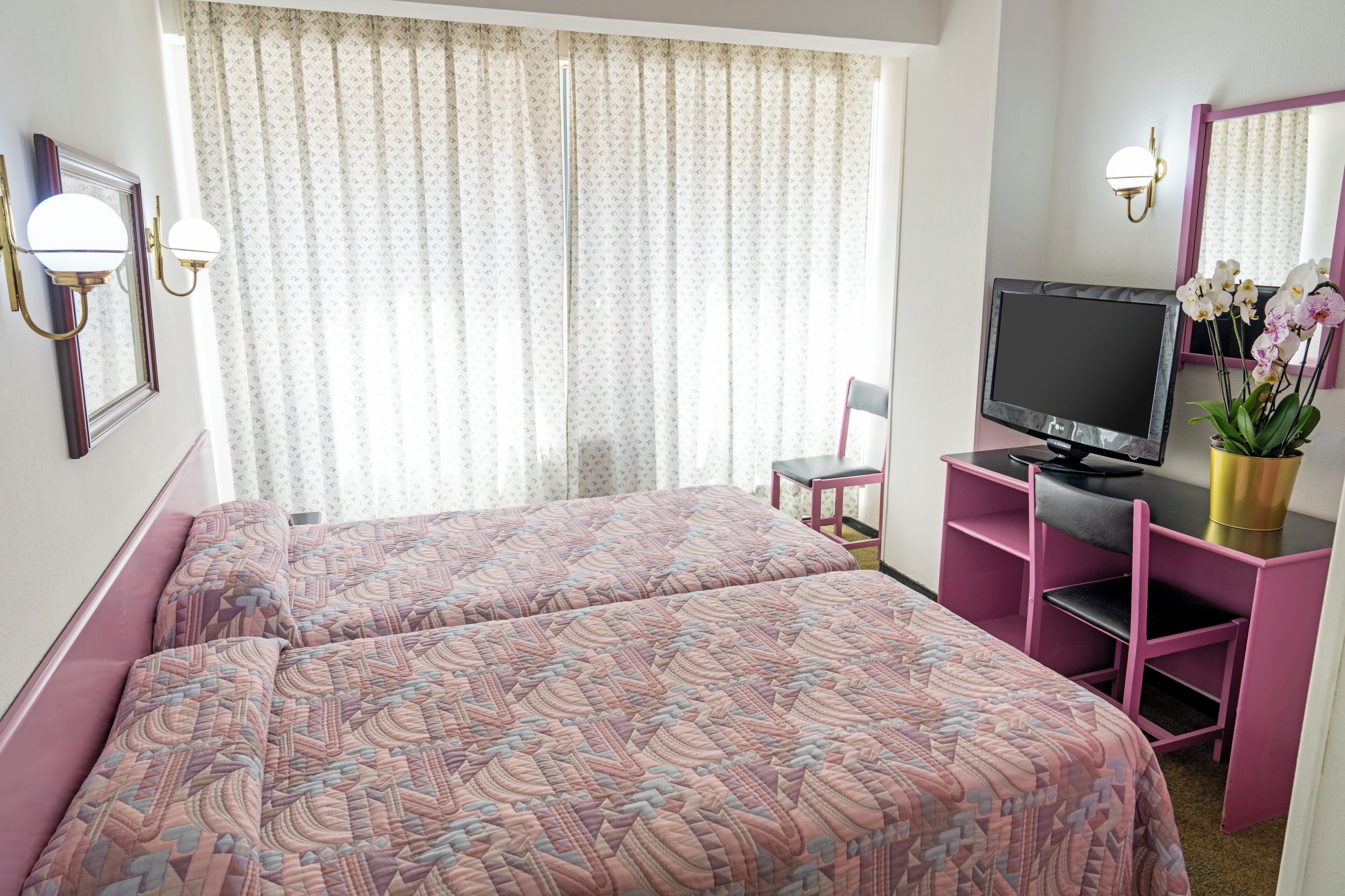 A hotel room with two beds, a TV, a desk, a chair, and a window with curtains.