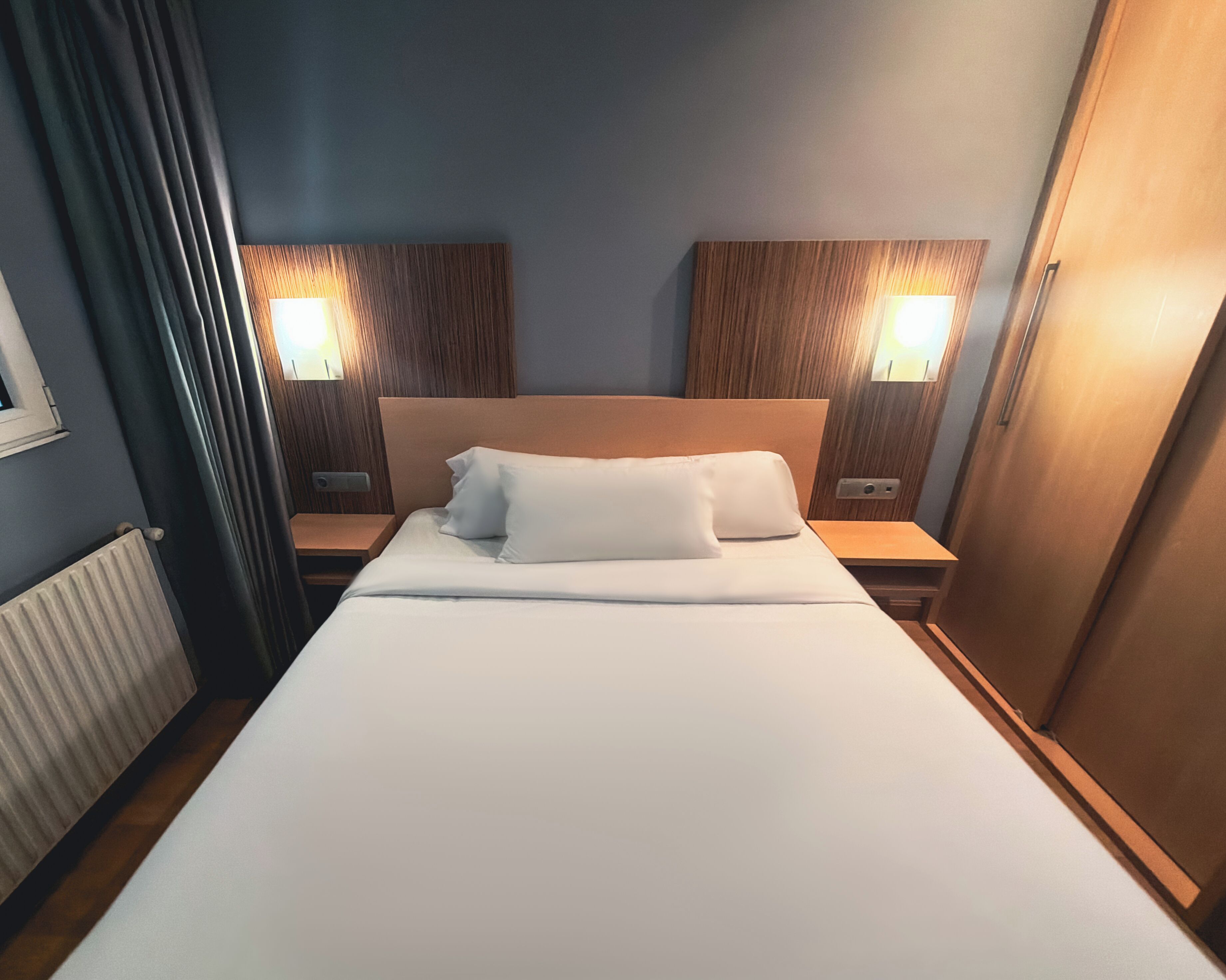 A hotel room with a bed, two bedside tables, a window with curtains, and two wall-mounted lamps.