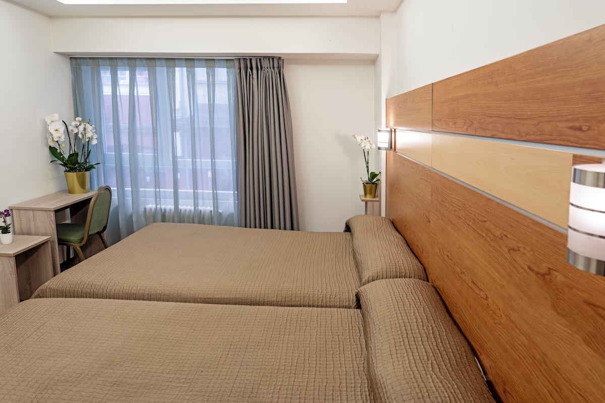 A hotel room with two beds, a desk, a chair, a wooden headboard, and a window with curtains.