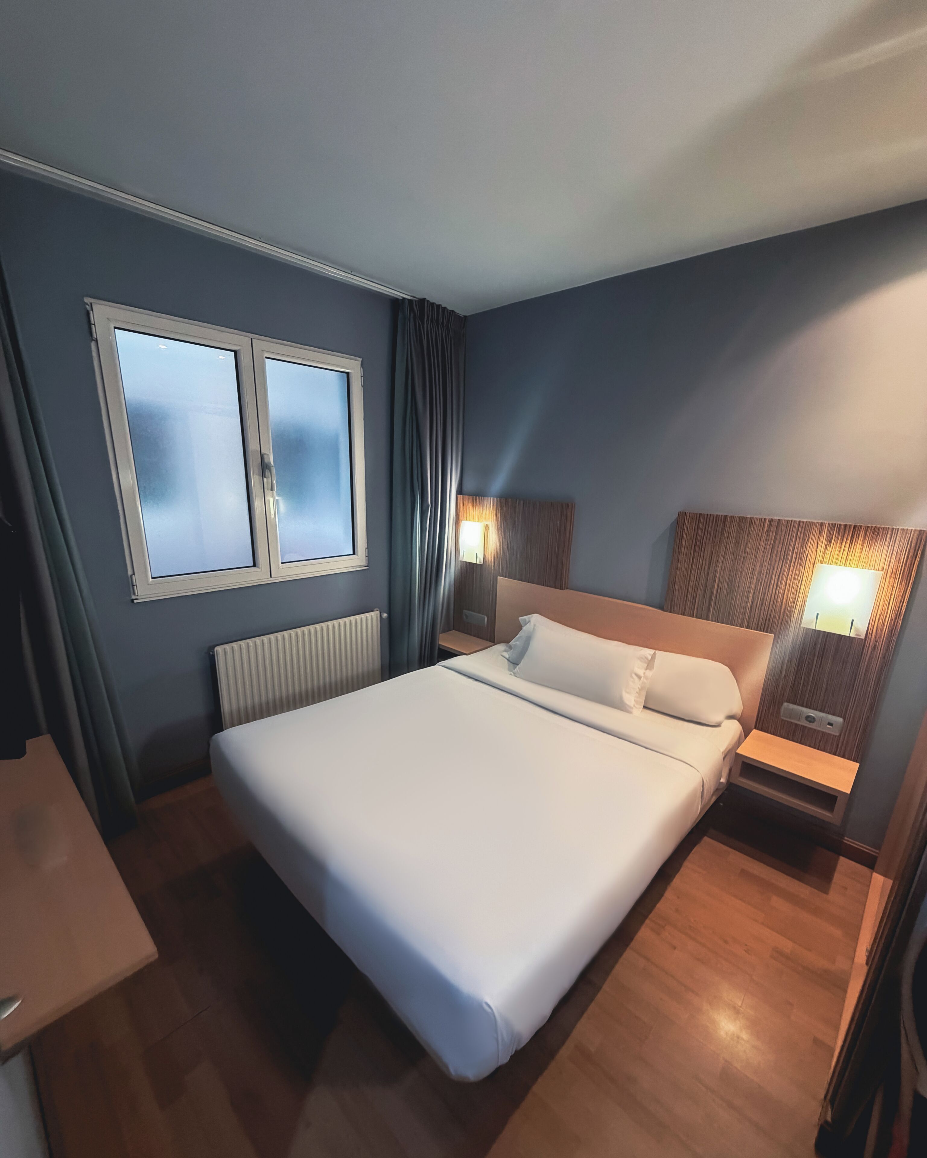 Una habitación de hotel con una cama grande, un escritorio, una ventana con cortinas, un cabecero con luz y una mesita de noche con lámpara.