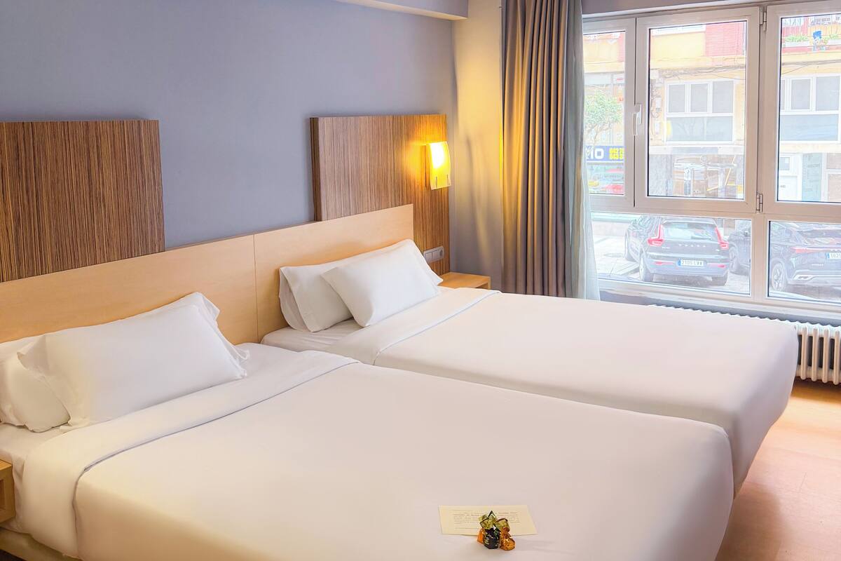 A hotel room with two beds, a nightstand, a wall-mounted lamp, and a window with curtains.