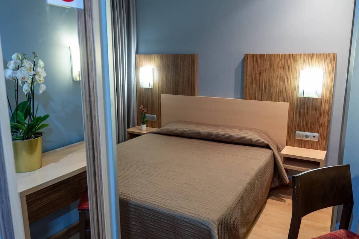 A hotel room with a bed, bedside tables, a desk, a chair, and a potted plant.