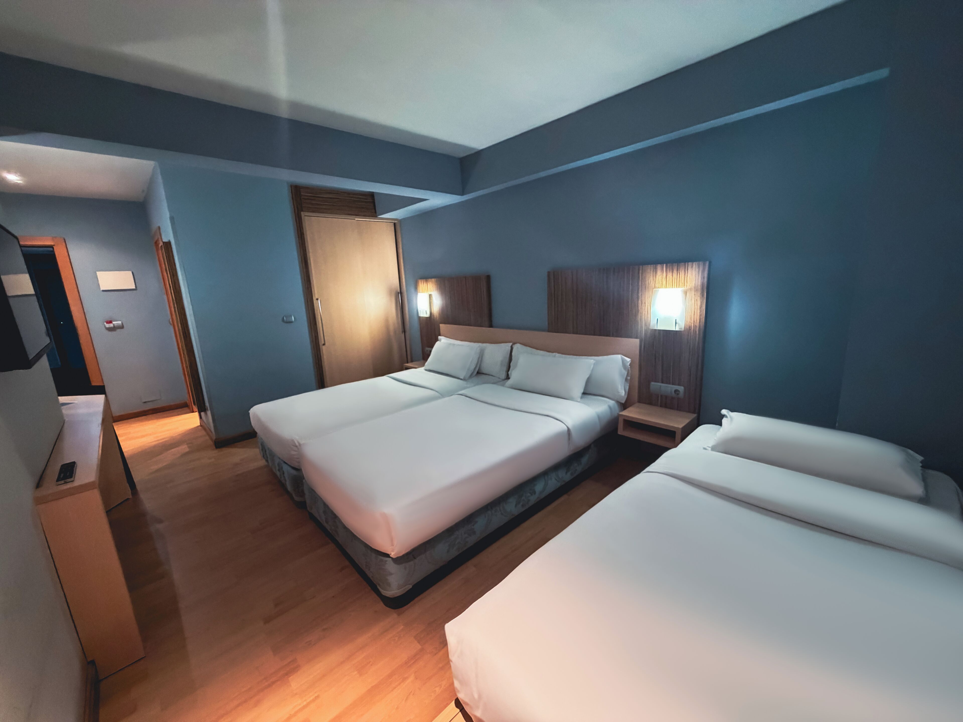 A hotel room with two beds, a wooden floor, a wardrobe, and a nightstand with a lamp.