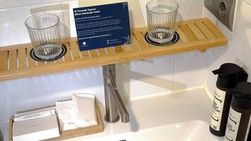 Un baño con un estante de madera que sostiene dos vasos de vidrio, un cartel sobre el compromiso ambiental y artículos de aseo personal, incluyendo una botella de jabón y una caja de pañuelos.