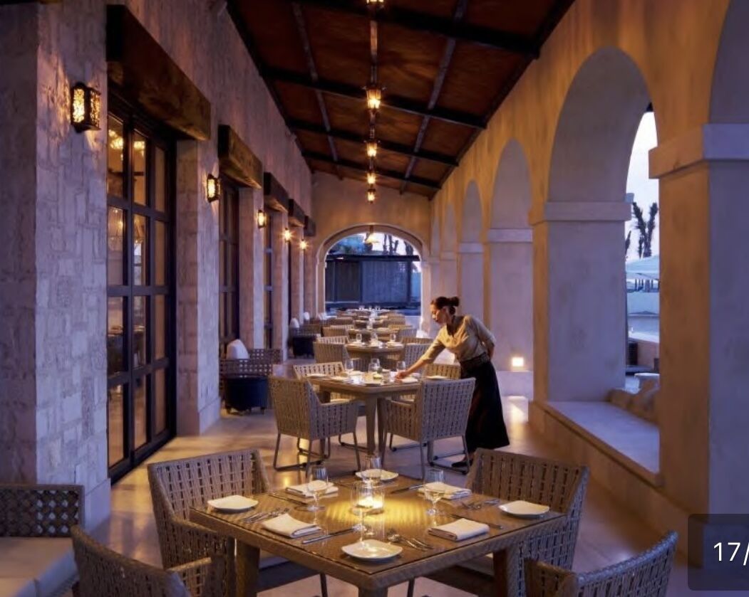 A restaurant with tables set for dining, a waiter attending to guests, and a view of the outdoors through an arched window.