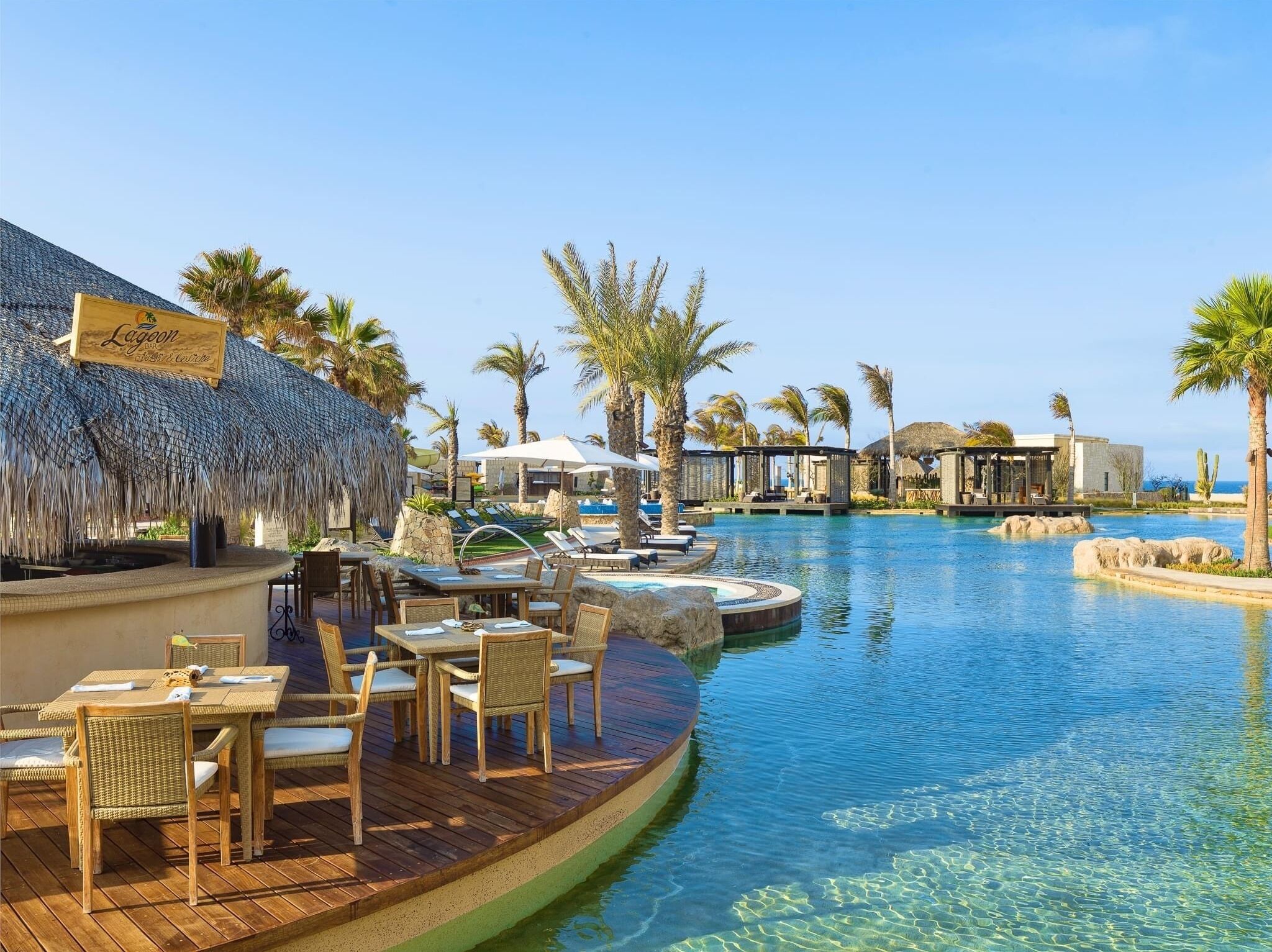 Un área de piscina en un resort con sillones de descanso, un bar y estructuras de techo de paja.