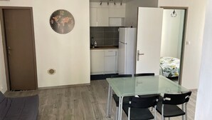 Un espace de vie compact comprenant un coin cuisine, un coin repas avec une table en verre et un canapé gris.