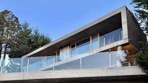 Ein modernes, zweistöckiges Haus mit großen Glasfenstern, einer Holzterrasse und Sitzgelegenheiten im Freien.