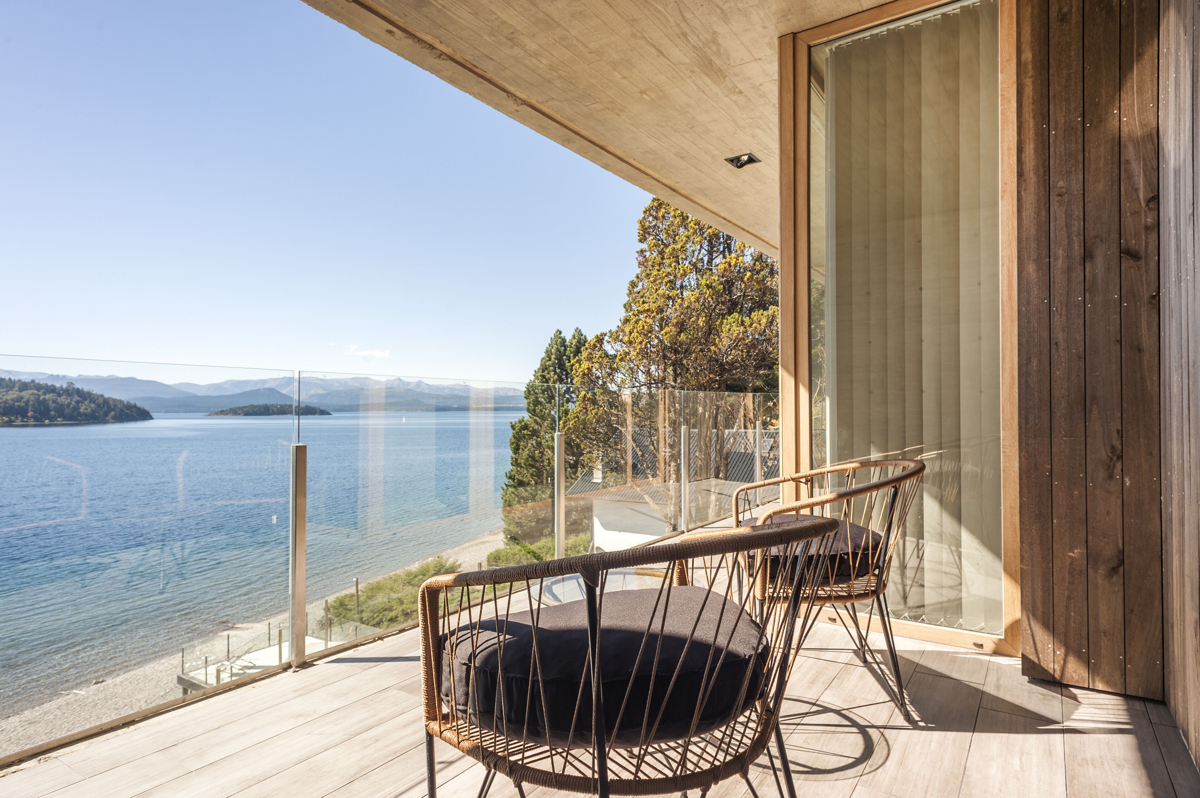 A balcony with wicker chairs, a glass railing, and a view of a lake and mountains.