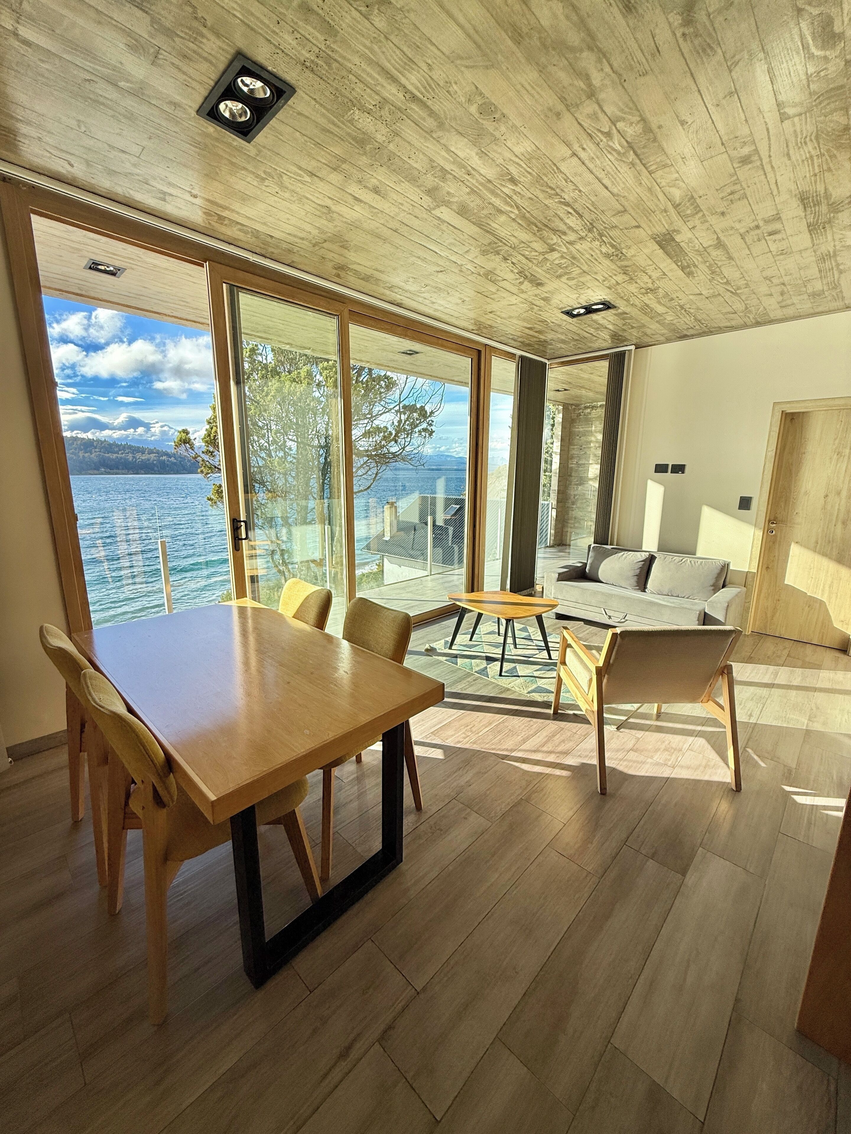 A modern interior with a wooden table and chairs, a sofa, and large windows offering a view of a lake and mountains.