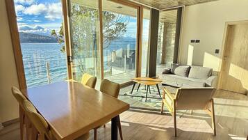 A modern interior with a wooden table and chairs, a sofa, and large windows offering a view of a lake and mountains.