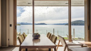 Ein Essbereich mit einem großen Tisch und Stühlen, einer Obstschale und Blick auf einen See mit Bergen im Hintergrund.
