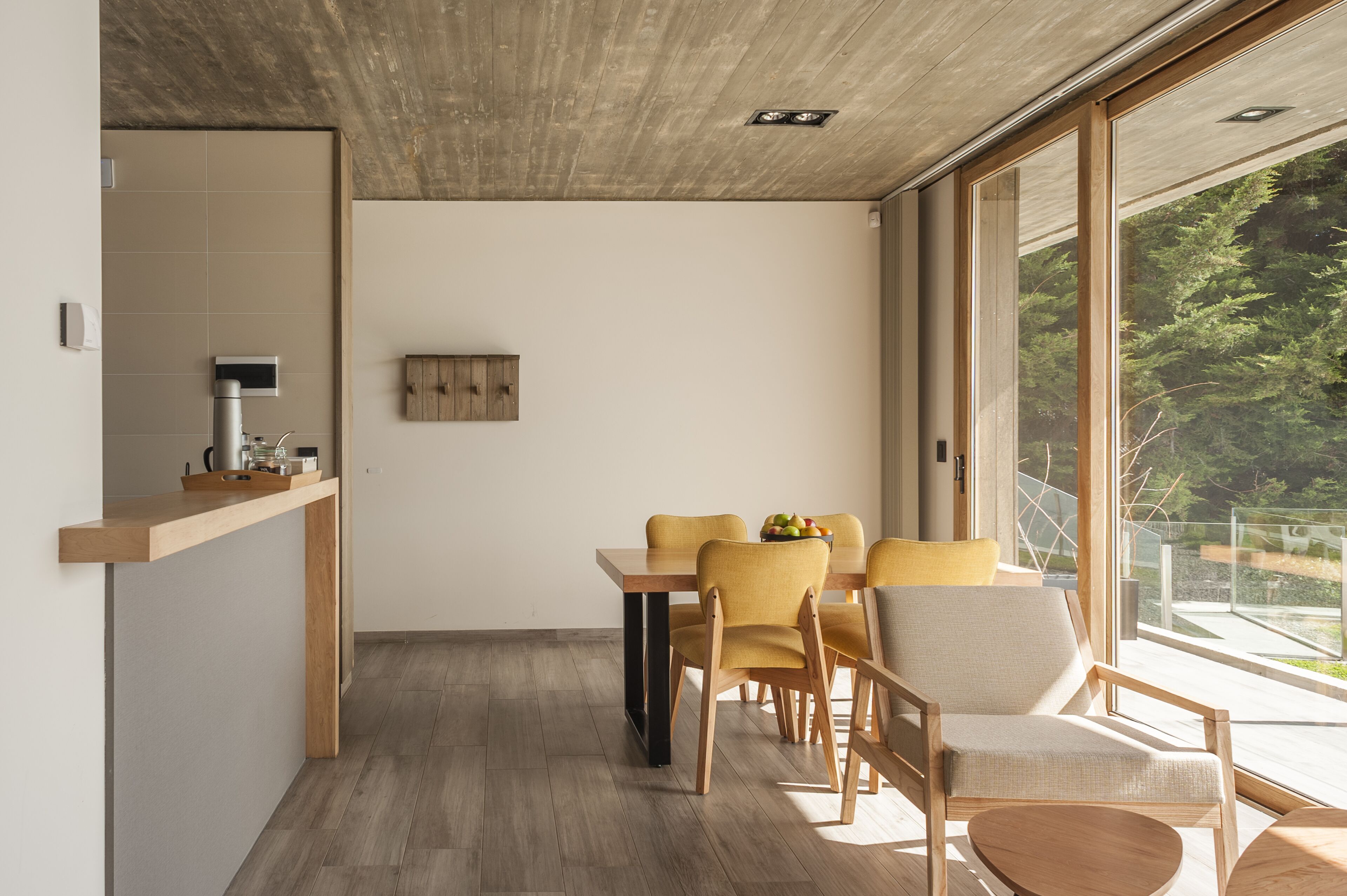 Un comedor moderno con una mesa y sillas de madera, una pequeña zona de cocina con una cafetera, y ventanas grandes que ofrecen vistas a zonas verdes.