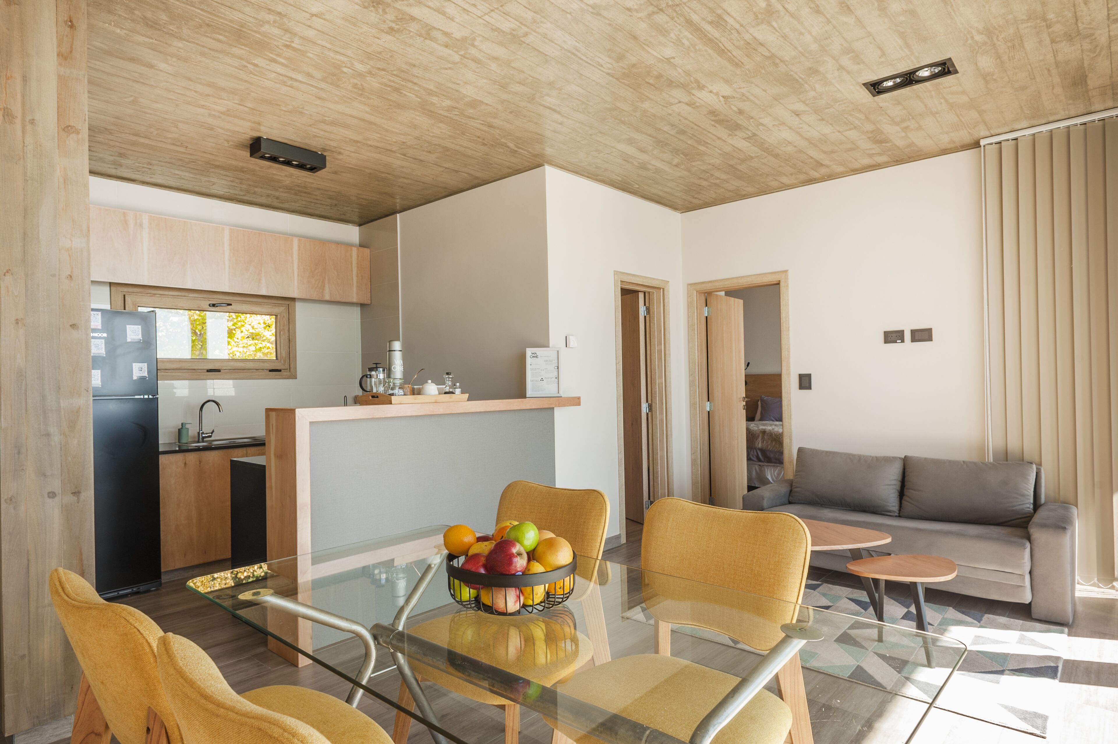 Een moderne leefruimte met een glazen eettafel, gele stoelen, een grijze bank en een keuken met houten kasten en een zwarte koelkast.