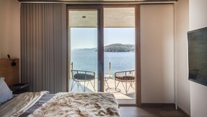 Ein Schlafzimmer mit einer großen Schiebeglastür, die auf einen Balkon mit zwei Stühlen und einem Tisch führt, mit Blick auf ein Gewässer und ein entferntes Land.