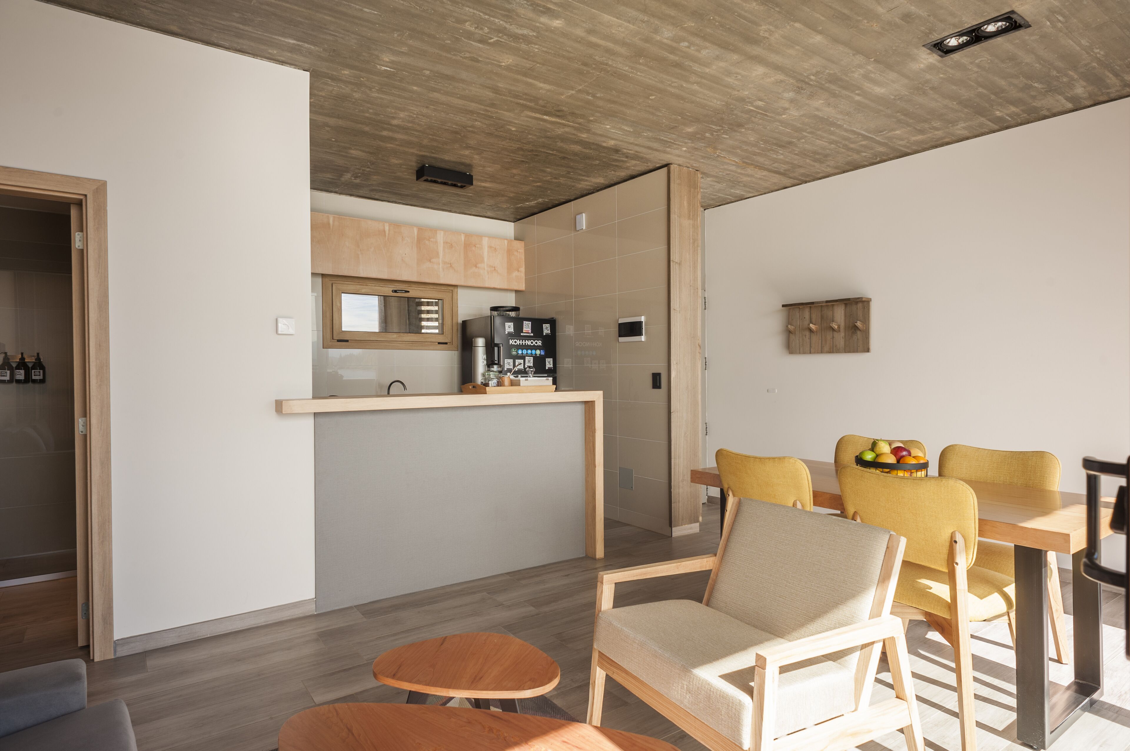 A modern kitchen with wooden cabinets, a built-in coffee machine, and a dining area with yellow chairs and a wooden table.