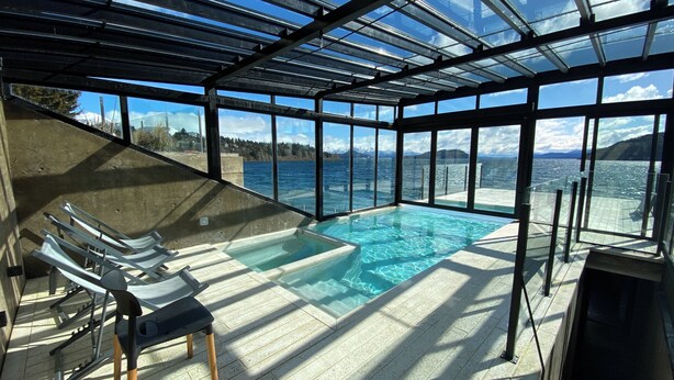 Ein von Glas umschlossener Infinity-Pool mit klarem Wasser, umgeben von einer Betonplattform und Liegestühlen.