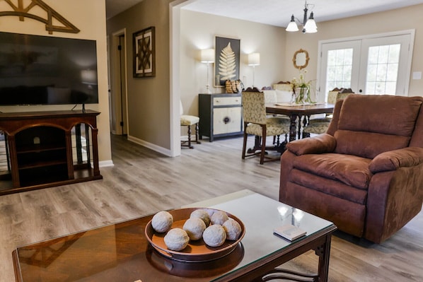 A living room with a brown leather armchair, a glass coffee table with a bowl of cookies, a television, and a dining area in the background.