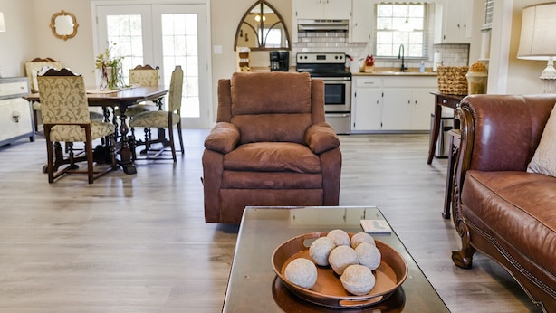 A spacious living room with a brown leather armchair, a dining area with patterned chairs, and a kitchen with white cabinets. - The McClellan Home w/King suite! (Anniston)