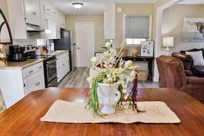 A cozy living room with a wooden dining table, a white vase with flowers, a brown leather sofa, and a kitchen in the background.