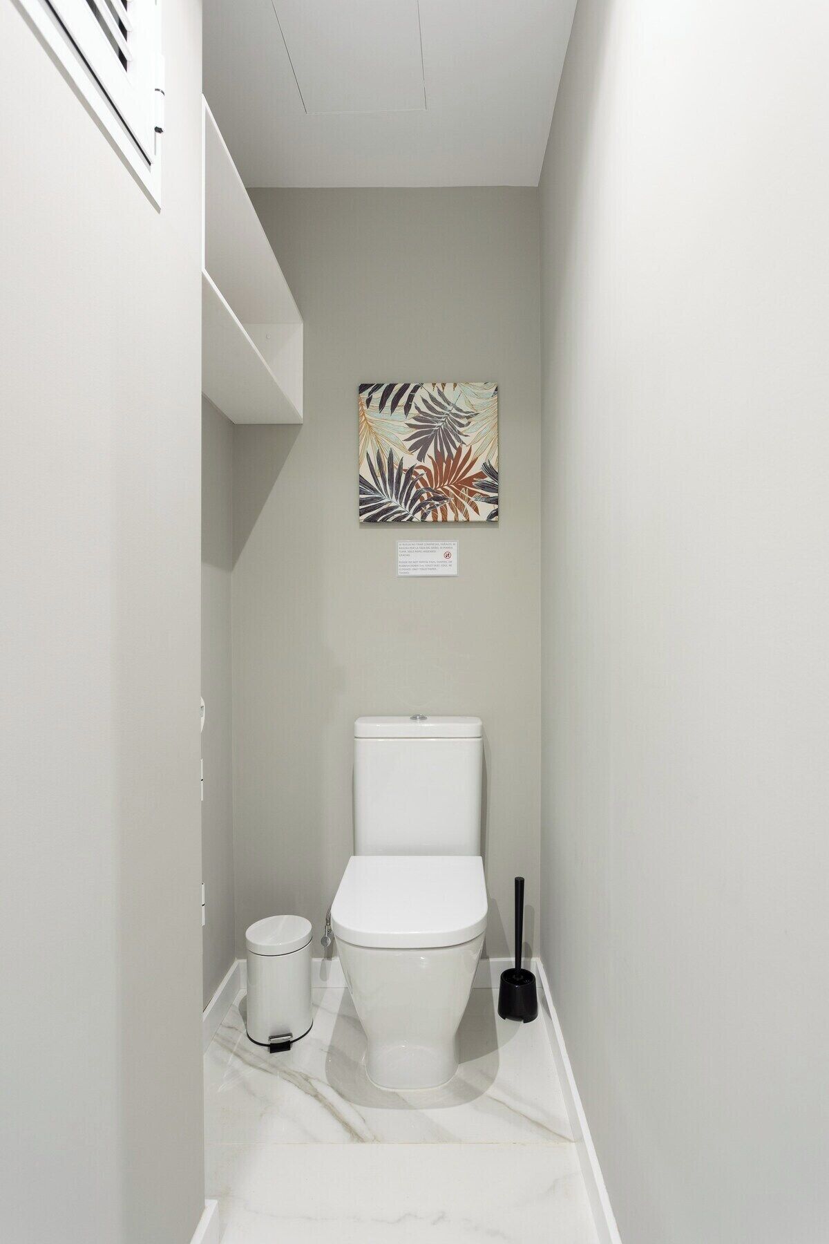 A small, narrow bathroom with a toilet, a trash bin, and a plunger.